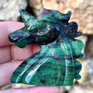 May include: A carved green and black unicorn figurine with red flecks. The sculpture is held in a hand, showcasing the details of the polished stone. The unicorn has a detailed mane and horn, with a smooth, glossy finish.