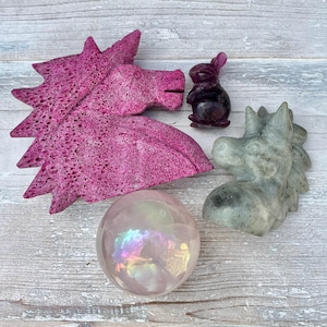 May include: A collection of carved stone figurines, including a pink unicorn head, a gray unicorn head, a purple rabbit, and a clear iridescent sphere. The objects are arranged on a light wood-grain surface.