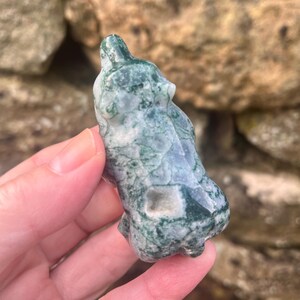 Puede incluir: Una talla de &aacute;gata musgosa pulida con forma de peque&ntilde;o animal, que presenta una base blanca transl&uacute;cida con inclusiones de color verde oscuro similares a helechos y patrones dendr&iacute;ticos en toda la superficie, con una textura suave y brillante.