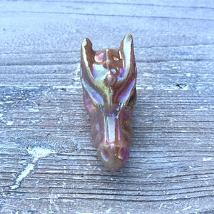 K&ouml;nnte beinhalten: Keramik-Drachenkopf-Figur mit schillerndem Finish. Der Drachenkopf hat eine hellbraune Farbe mit rosa und goldenen Akzenten. Die Figur liegt auf einer verwitterten Holzoberfl&auml;che.