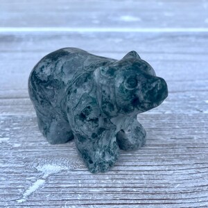 Puede incluir: Una peque&ntilde;a figura de oso tallada a mano en piedra de color verde oscuro con vetas. La escultura presenta una superficie lisa y pulida con variaciones de color y vetas naturales visibles en todo el cuerpo, representando a un oso de pie.