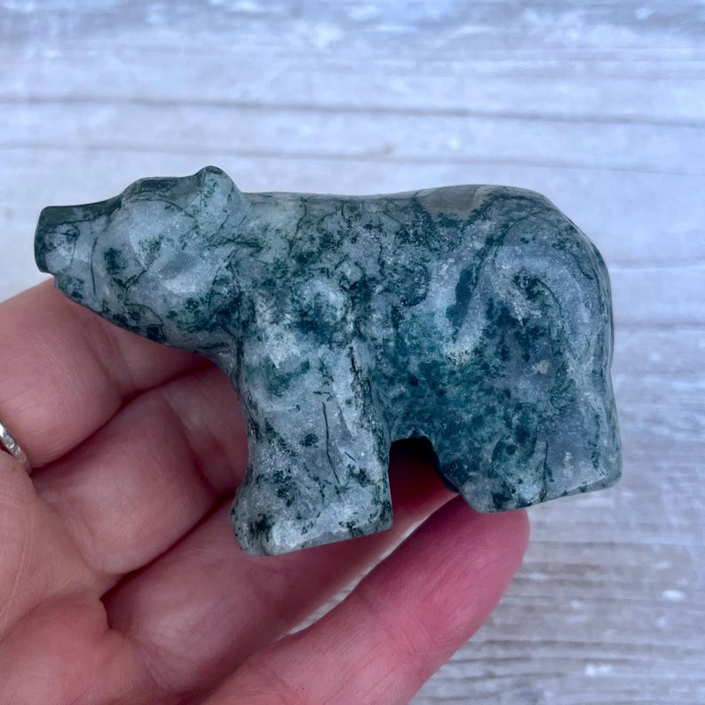 Puede incluir: Una peque&ntilde;a figura de oso tallada a mano en &aacute;gata musgosa. La piedra presenta una base gris p&aacute;lido transl&uacute;cida con inclusiones de color verde bosque similares al musgo y vetas naturales en toda la superficie lisa y pulida del animal.