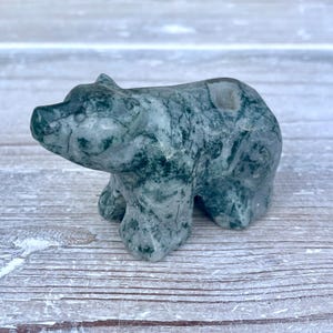 Puede incluir: Una peque&ntilde;a figura de oso tallada a mano en piedra de &aacute;gata musgosa pulida. La escultura presenta una apariencia moteada y semitransl&uacute;cida con inclusiones de color verde bosque profundo y vetas gris&aacute;ceas blanquecinas en la superficie.