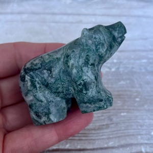 Puede incluir: Una peque&ntilde;a figura de oso tallada a mano en &aacute;gata musgosa. La piedra presenta un patr&oacute;n jaspeado con inclusiones de color verde bosque profundo y blanco transl&uacute;cido, mostrando una superficie lisa y pulida y una forma de oso estilizada.