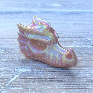 K&ouml;nnte beinhalten: Kleine, schillernde Drachenkopf-Figur in Rosa-, Gold- und Lilat&ouml;nen. Die Skulptur weist detaillierte Merkmale wie Schuppen und H&ouml;rner auf. Der Drache ist ein dekoratives Objekt mit Fantasy-Thema.