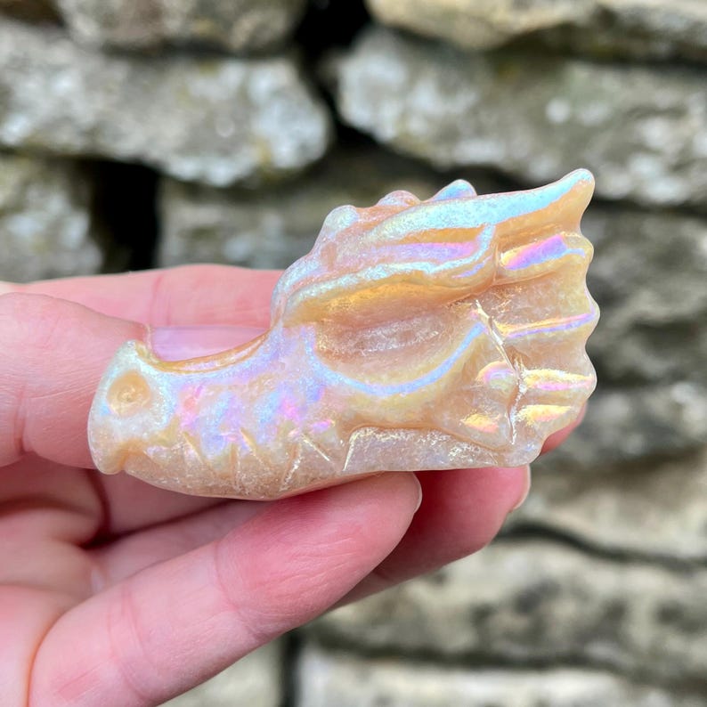 K&ouml;nnte beinhalten: Kleine, schillernde Drachenkopfskulptur. Die Skulptur wird in einer Hand gehalten und hat eine perlmuttartige Oberfl&auml;che mit Gold-, Rosa- und Blaut&ouml;nen. Der Drachenkopf hat detaillierte Merkmale, einschlie&szlig;lich Schuppen und H&ouml;rner, und besteht aus einem durchscheinenden Material.