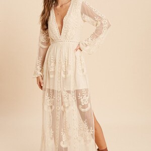 May include: A long, white, lace maxi dress with a plunging neckline and long sleeves. The dress has a fitted bodice and a flowing skirt with a side slit. The dress is made of delicate lace and has a sheer, ethereal look.