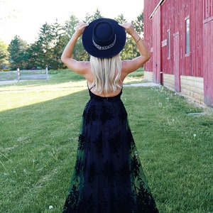 May include: A woman wearing a black maxi dress with floral embroidery and a black wide-brimmed hat. She is standing in a grassy field with a red barn in the background.