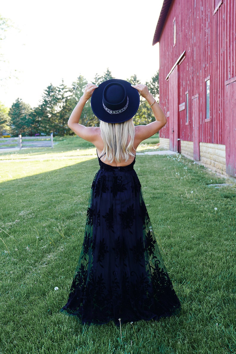 May include: A woman wearing a black maxi dress with floral embroidery and a black wide-brimmed hat. She is standing in a grassy field with a red barn in the background.