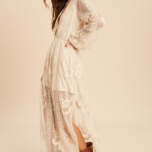 May include: A long, white, lace maxi dress with a plunging neckline and long sleeves. The dress has a fitted bodice and a flowing skirt. The model is wearing brown ankle boots.