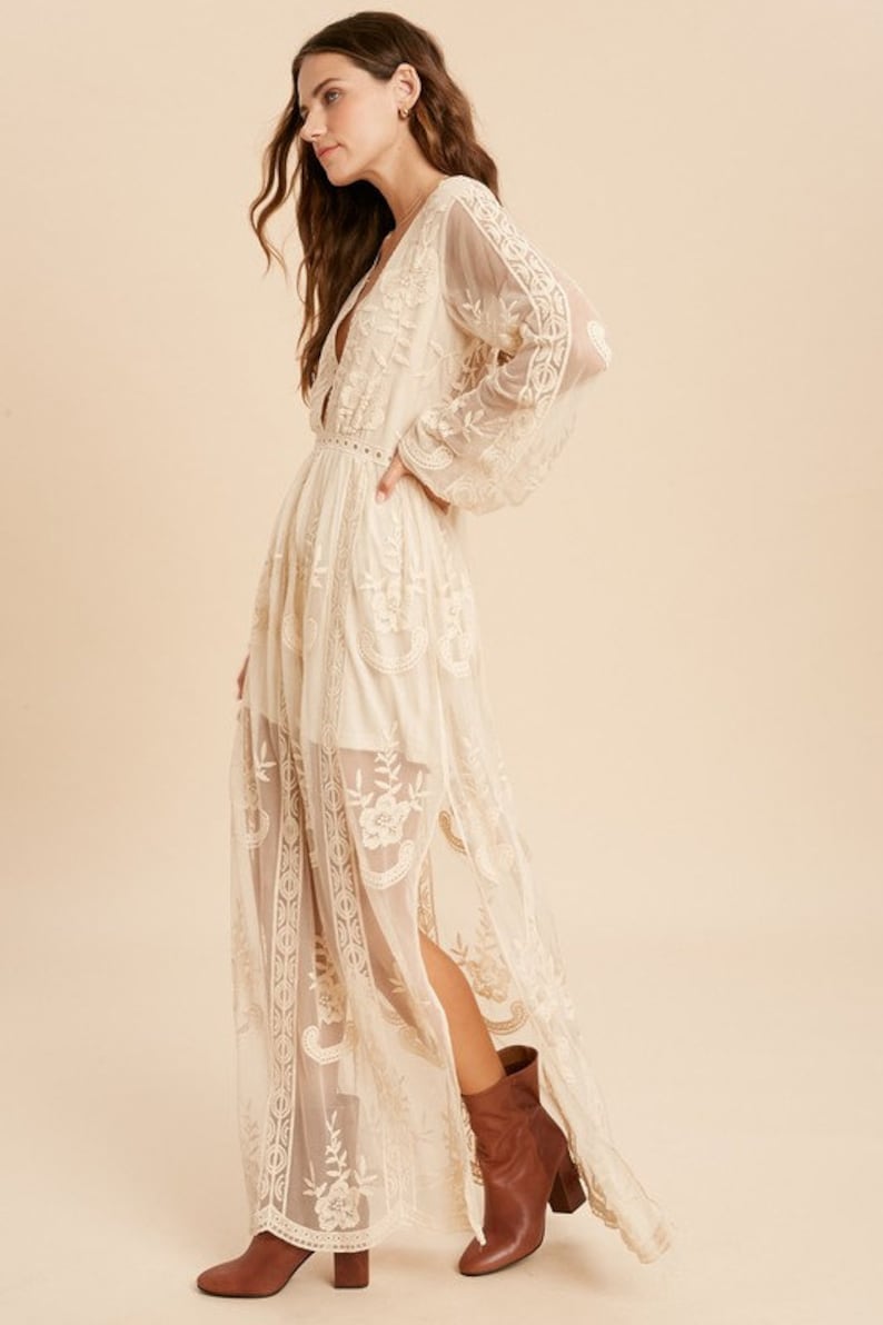 May include: A long, white, lace maxi dress with a plunging neckline and long sleeves. The dress has a fitted bodice and a flowing skirt. The model is wearing brown ankle boots.