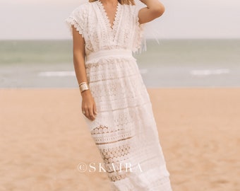 May include: A woman wearing a white crochet maxi dress with a wide-brimmed straw hat. The dress has a V-neckline and short sleeves. The woman is standing on a sandy beach.