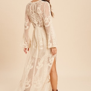 May include: A white maxi dress with a lace overlay and a side slit. The dress has a V-neckline and long sleeves.