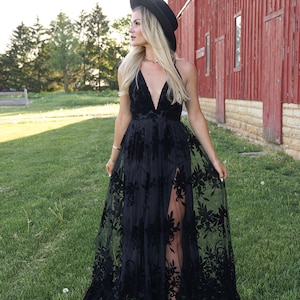 May include: A woman wearing a black maxi dress with floral embroidery and a black wide-brimmed hat. She is standing in a field with a red barn in the background.