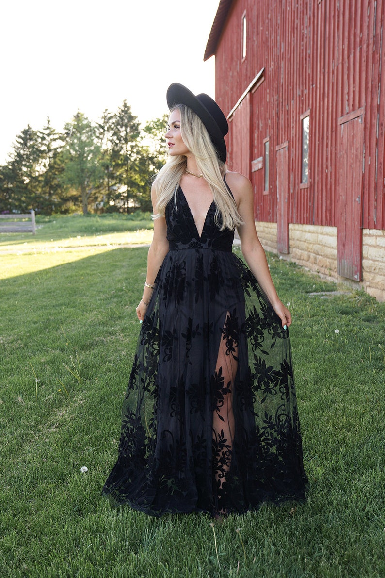 May include: A woman wearing a black maxi dress with floral embroidery and a black wide-brimmed hat. She is standing in a field with a red barn in the background.