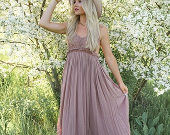 May include: A woman wearing a light brown maxi dress with a lace bodice and a wide-brimmed hat, standing in a field of green grass and white flowering trees.