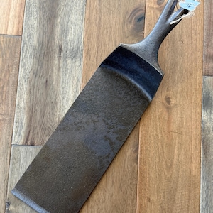 May include: A dark gray cast iron spatula with a long, flat blade and a handle with a loop for hanging. The spatula has a textured surface and a slightly curved blade edge. A small tag is attached to the handle.