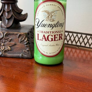 May include: A green candle in a repurposed Yuengling Lager beer bottle. The label features an eagle and the words "Yuengling Traditional Lager." The candle is filled with green wax and is 12 fl oz.