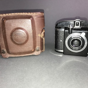 May include: A black vintage Beacon camera with a silver lens and a brown leather case. The camera has a silver knob on the top and a silver trim around the lens. The case has the word "Beacon" embossed on the front.