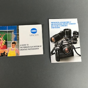 May include: Two Minolta camera instruction booklets. The first booklet has a blue and white logo and the text "A Guide to the Minolta SLR System of Creative Photography". The second booklet has a black and white photo of a Minolta camera with a lens attached and the text "Minolta Answers 7 Questions Most Asked By New Camera Owners."