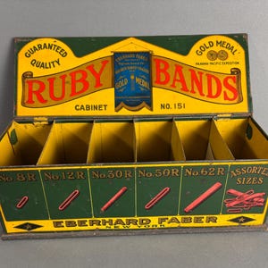 May include: A vintage tin box with compartments for storing rubber bands. The box is green and yellow with the words "Ruby Bands" in red and gold lettering. The box is labeled "No. 151 Cabinet" and features a gold medal logo.