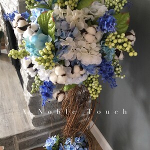 Könnte beinhalten: Ein weißer Topf mit einer braunen geflochtenen Basis ist mit einem Blumenarrangement aus blauen und weißen Hortensien, Baumwollknäueln und grünen Beeren gefüllt. Der Text "A Noble Touch" ist auf dem Topf sichtbar.