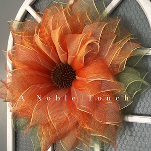 May include: A large, orange and green mesh sunflower wreath with a brown center. The wreath is hanging on a white door with a wire mesh window behind it. The text "A Noble Touch" is visible in the bottom center of the image.