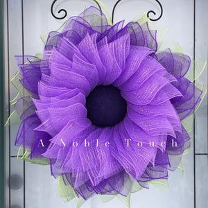 Könnte beinhalten: Ein lila-grüner Mesh-Kranz in Form einer Blume. Der Kranz hat eine dunkelviolette Mitte und ist mit Glitzer verziert. Der Text "A Noble Touch" ist in der Mitte unten am Kranz sichtbar.