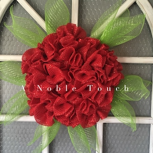 May include: A red and green wreath made of mesh with a glittery finish. The wreath is decorated with green mesh leaves. The wreath is hanging on a white door with a wire mesh backing.