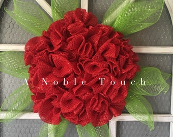 May include: A red and green wreath made of mesh with a glittery finish. The wreath is decorated with green mesh leaves. The wreath is hanging on a white door with a wire mesh backing.