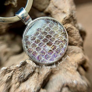 Op de afbeelding: Zilverkleurige sleutelhanger met een ronde hanger. De hanger heeft een getextureerd, iriserend ontwerp in paarse, gouden en bruine tinten, gezet in een heldere laag. De sleutelhanger rust op een stuk hout.