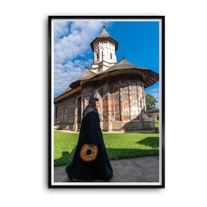 May include: A person in a long black robe walks past an Eastern Orthodox church with exterior wall frescoes. The person carries a round, golden-brown bread loaf. The church features a tall central tower and a dark roof against a blue sky.