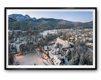 May include: An aerial view of a snow-covered village nestled in a valley surrounded by snow-capped mountains. The village is covered in a blanket of fresh snow, and the houses have snow-covered roofs. The trees are also covered in snow, and the sky is a clear blue.