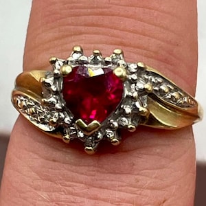 May include: A gold ring featuring a central heart-shaped red gemstone surrounded by a halo of small clear stones. The band is crafted from yellow gold and includes additional small clear stones set along the shoulders of the ring shank.
