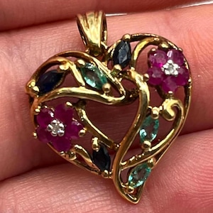 May include: A gold heart-shaped pendant featuring an openwork design with two five-petaled magenta flowers, each with a small white center stone. The piece is accented with marquise-cut dark blue sapphires and light green emerald-colored stones.