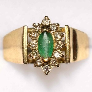 May include: A gold ring featuring a marquise-cut emerald center stone surrounded by a halo of small diamonds. The ring has a wide, flat band and a decorative setting. The emerald is a vibrant green, and the diamonds sparkle against the gold.