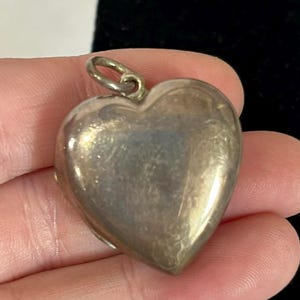 May include: A silver heart-shaped pendant with a small loop at the top. The pendant has a smooth, reflective surface and appears to be made of metal. The heart is slightly rounded and has a vintage or antique appearance.