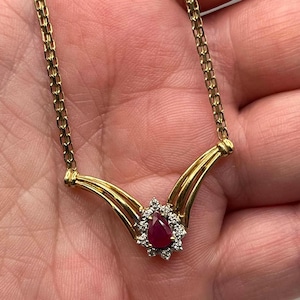 May include: A gold chain necklace featuring a V-shaped pendant with a central pear-shaped red ruby gemstone surrounded by a halo of small white diamonds. The metal has a polished finish, and the chain consists of interconnected rectangular links.