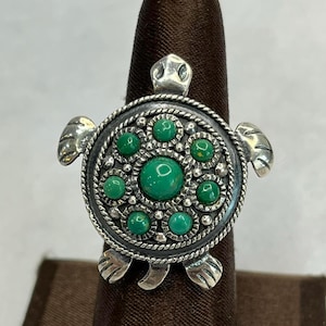 May include: A silver-toned ring in the shape of a turtle, featuring a circular shell design with a rope-like border. The shell is adorned with multiple round, bright green stones. The turtle's head, flippers, and feet are also visible.
