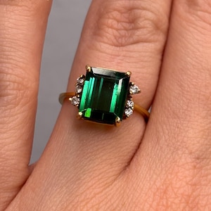 May include: A gold ring featuring a large, emerald-cut, deep green gemstone. The ring is accented with small, clear gemstones on either side of the main stone. The ring's band is a simple, polished gold.