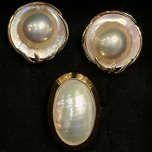 May include: A set of three pearl jewelry pieces, including a ring and two earrings. The ring has an oval-shaped pearl set in a gold band. The earrings are round and have a pearl set in a gold setting with a decorative design.