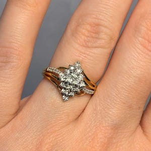 May include: A gold ring with a cluster of small diamonds in a floral design. The ring is on a person's finger, with dark red nail polish. The ring has a delicate band and a layered design.