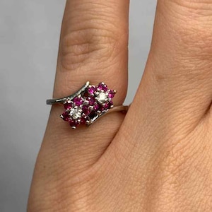 May include: A silver ring featuring a cluster of small, round, red gemstones and a single white gemstone. The gemstones are set in a floral-like arrangement. The ring is on a finger with a dark teal nail polish.