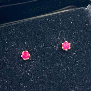 May include: A pair of round, vibrant pink stud earrings set in gold-toned metal prongs. The earrings are displayed inside a black velvet-lined jewelry box, which features a hinged lid and a soft, dark interior surface for secure storage.