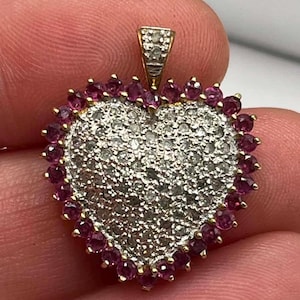 May include: A heart-shaped pendant featuring a center pavé of small clear stones surrounded by a border of vibrant pink stones. The piece is set in yellow gold and includes a small triangular bail at the top, also accented with tiny clear stones.