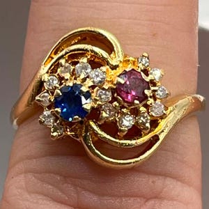 May include: A gold bypass ring featuring two central round-cut gemstones, one deep blue and one vibrant pink, each surrounded by a cluster of small clear white stones. The gold band has a swirling, fluid design that wraps around the central stones.