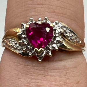 May include: A gold ring featuring a central heart-shaped deep pink gemstone. The stone is surrounded by small clear stones set in a silver-toned metal frame. The band transitions from a silver-toned setting at the center to smooth gold sides.