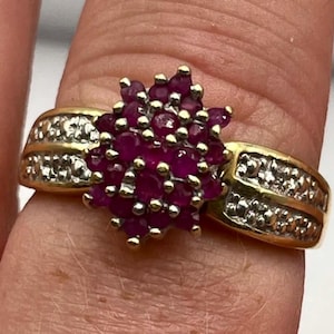 May include: A gold ring featuring a cluster of small, round, deep red rubies at the center. The band is crafted from yellow gold and is accented with a row of tiny, clear, sparkling diamonds embedded along each side of the central ruby cluster.