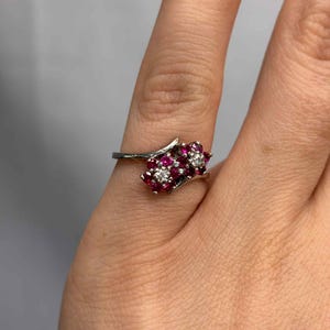 May include: A silver ring featuring clusters of red gemstones and a single cluster of white gemstones. The ring is set on a finger, showcasing its delicate design and intricate details. The gemstones are arranged in a floral pattern.