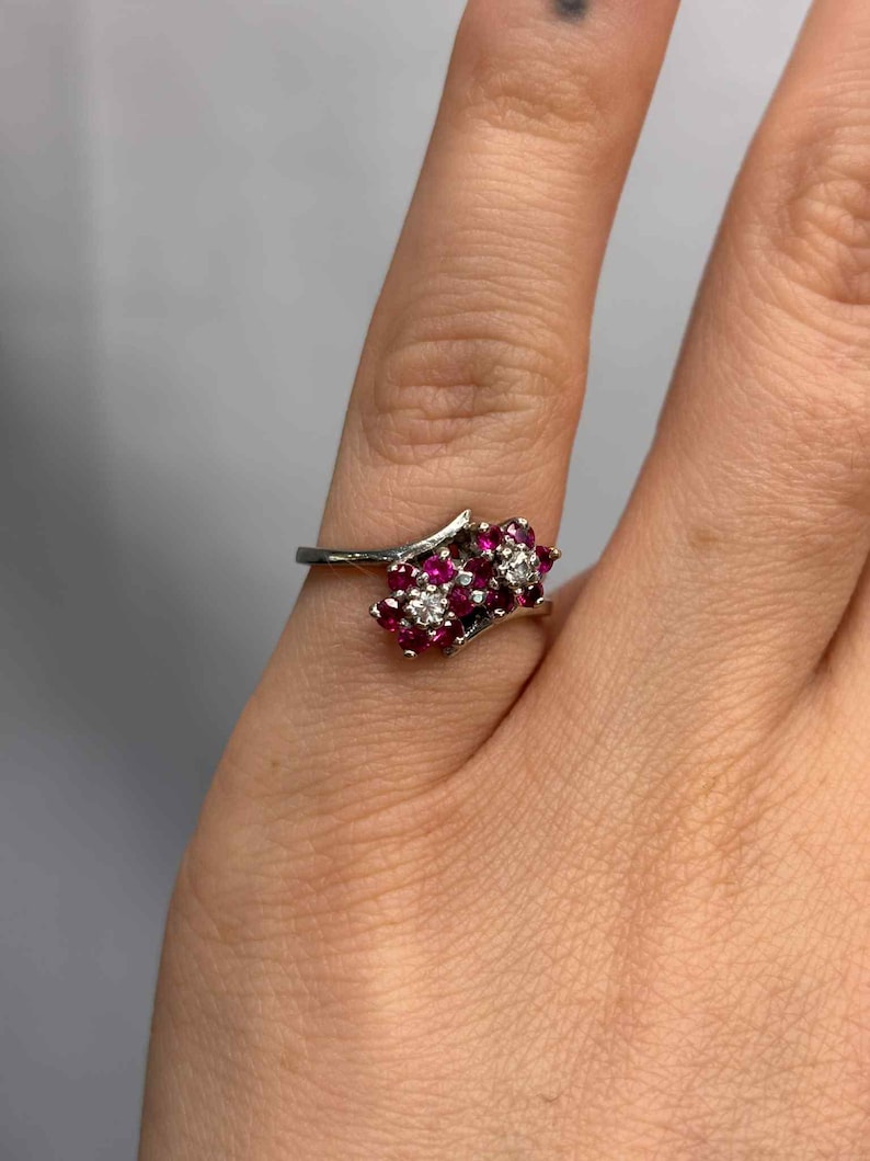 May include: A silver ring featuring clusters of red gemstones and a single cluster of white gemstones. The ring is set on a finger, showcasing its delicate design and intricate details. The gemstones are arranged in a floral pattern.